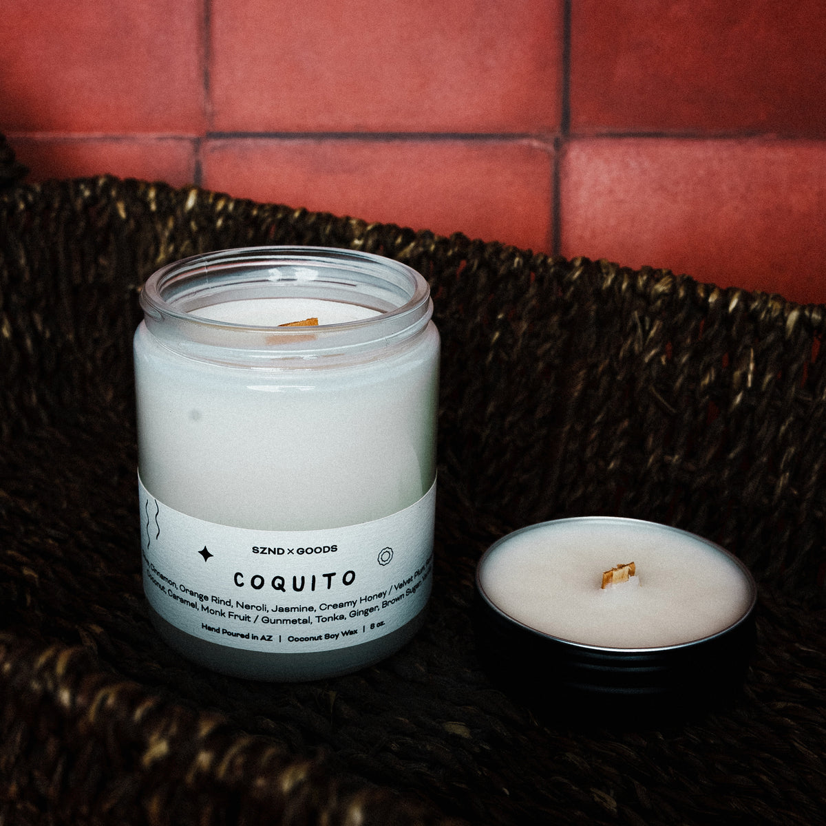 Candle labeled 'Coquito' on a woven surface with a red tiled wall background with it's matching mini 2oz candle.