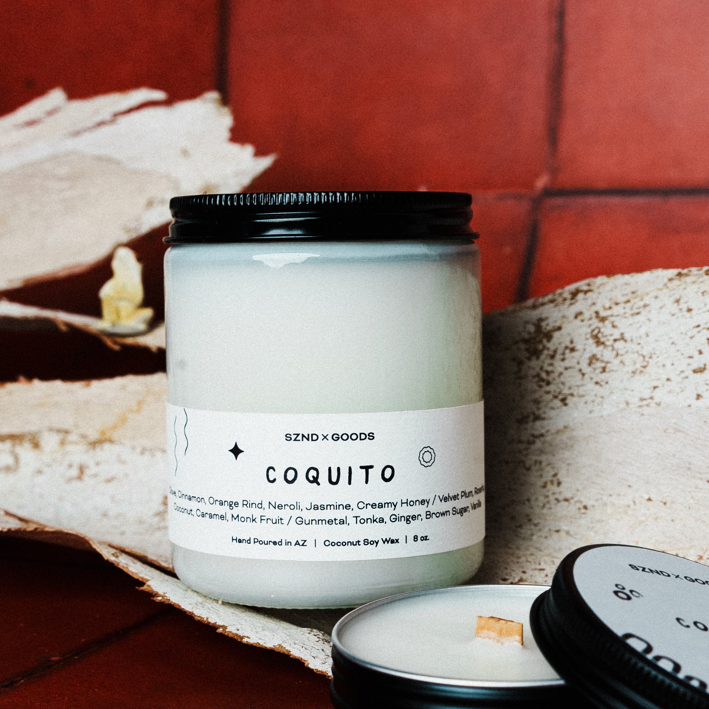 Candle jar labeled 'Coquito' by SZND x GOODS on a textured surface with a mini 2oz candle and coqui in the background.