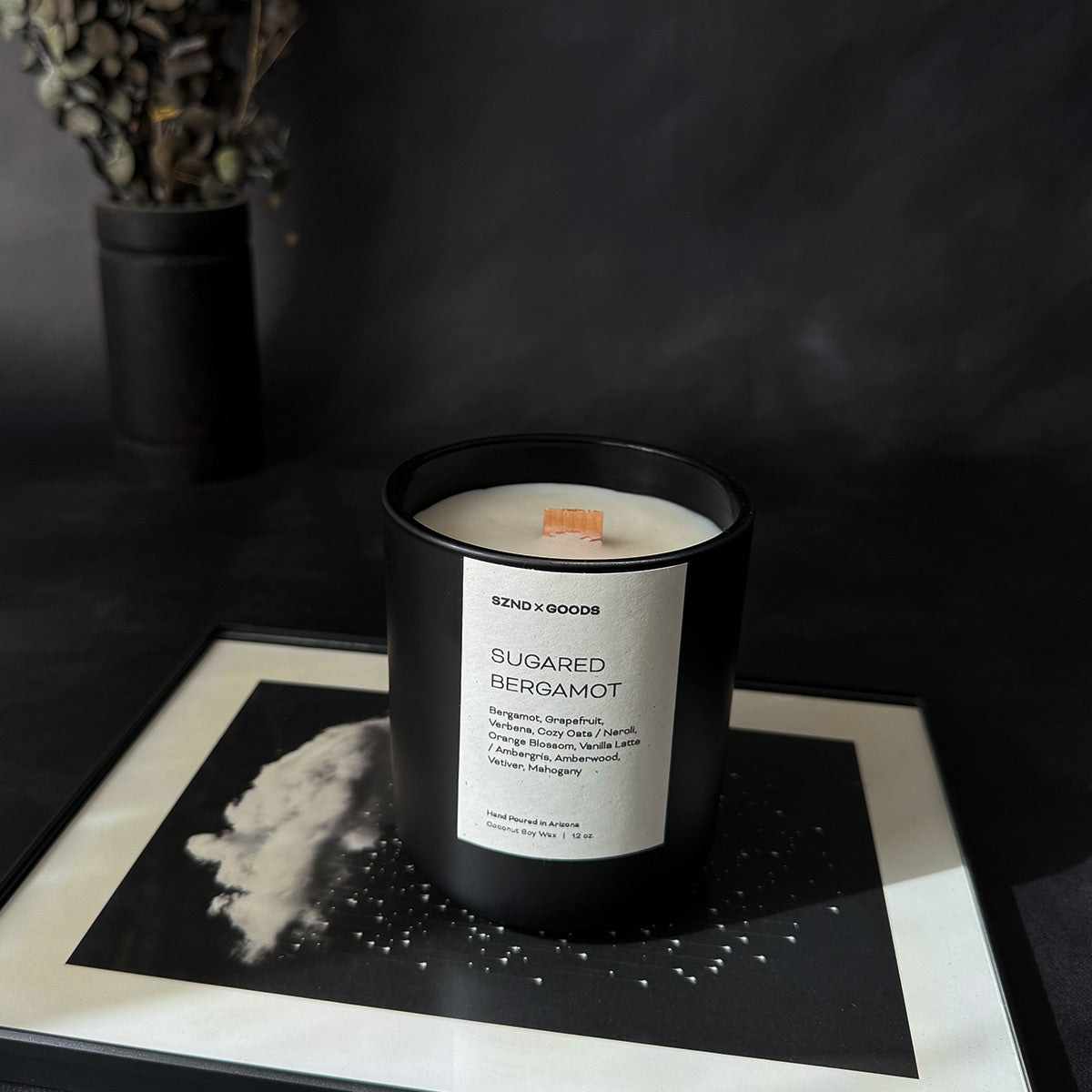 A black container candle with a wooden wick, labeled 'SUGARED BERGAMOT', with a white wax melt beside it, on a dark background.