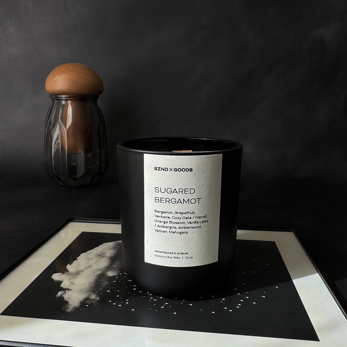 A black container candle with a wooden wick, labeled 'SUGARED BERGAMOT', with a white wax melt beside it, on a dark background.