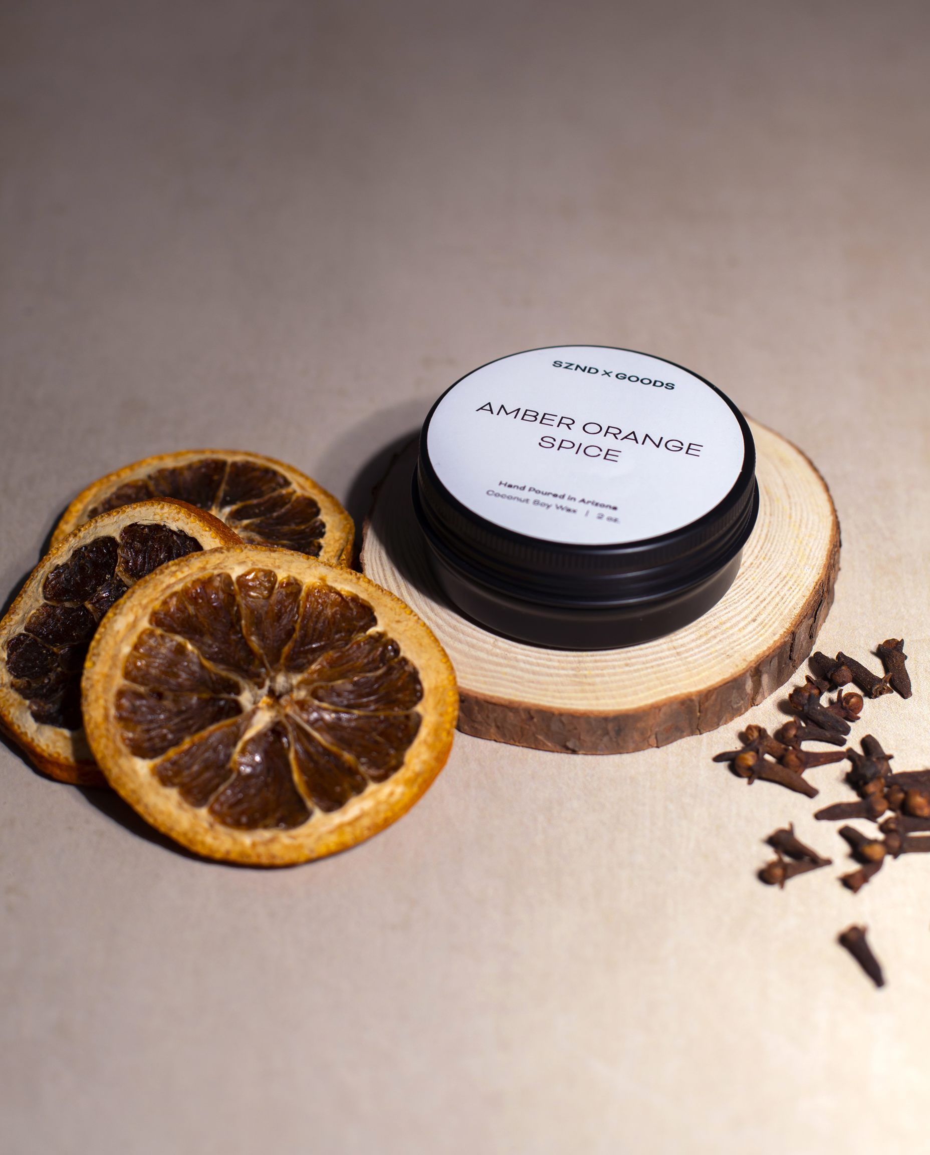 A black round mini candle labeled AMBER ORANGE SPICE sits on a wood slice, styled with dried orange slices and cloves on a neutral beige background.