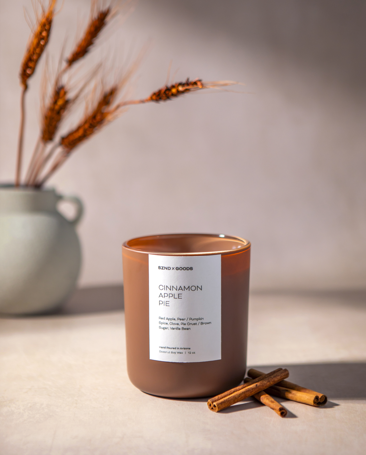 Cinnamon Apple Pie candle in a matte brown jar that sits on a light beige surface with cinnamon sticks and dried wheat in a vase behind it.