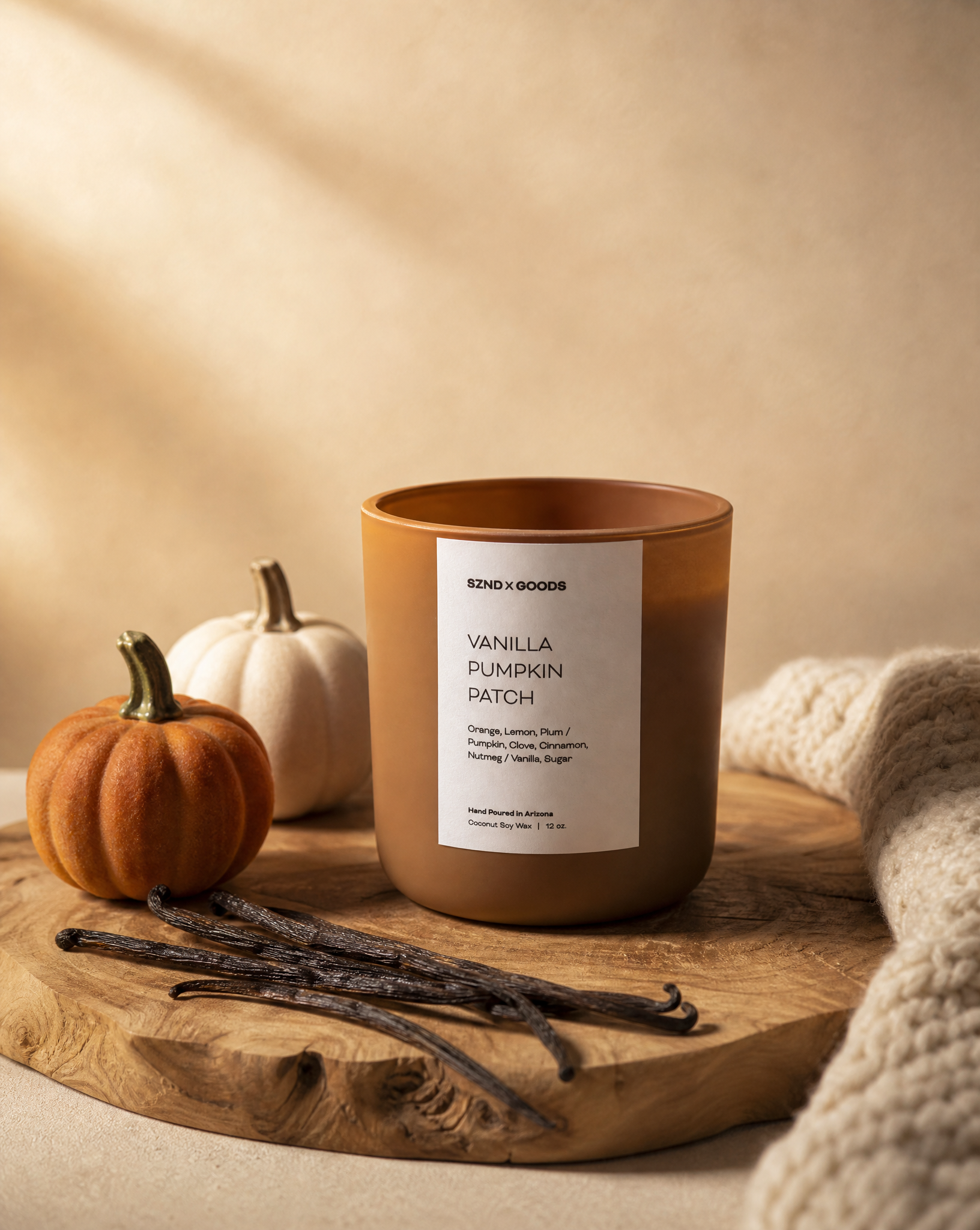The Vanilla Pumpkin Patch candle in a matte brown jar sits on a rustic wooden board, styled with vanilla beans, a small orange pumpkin, a white pumpkin, and a chunky knit blanket.