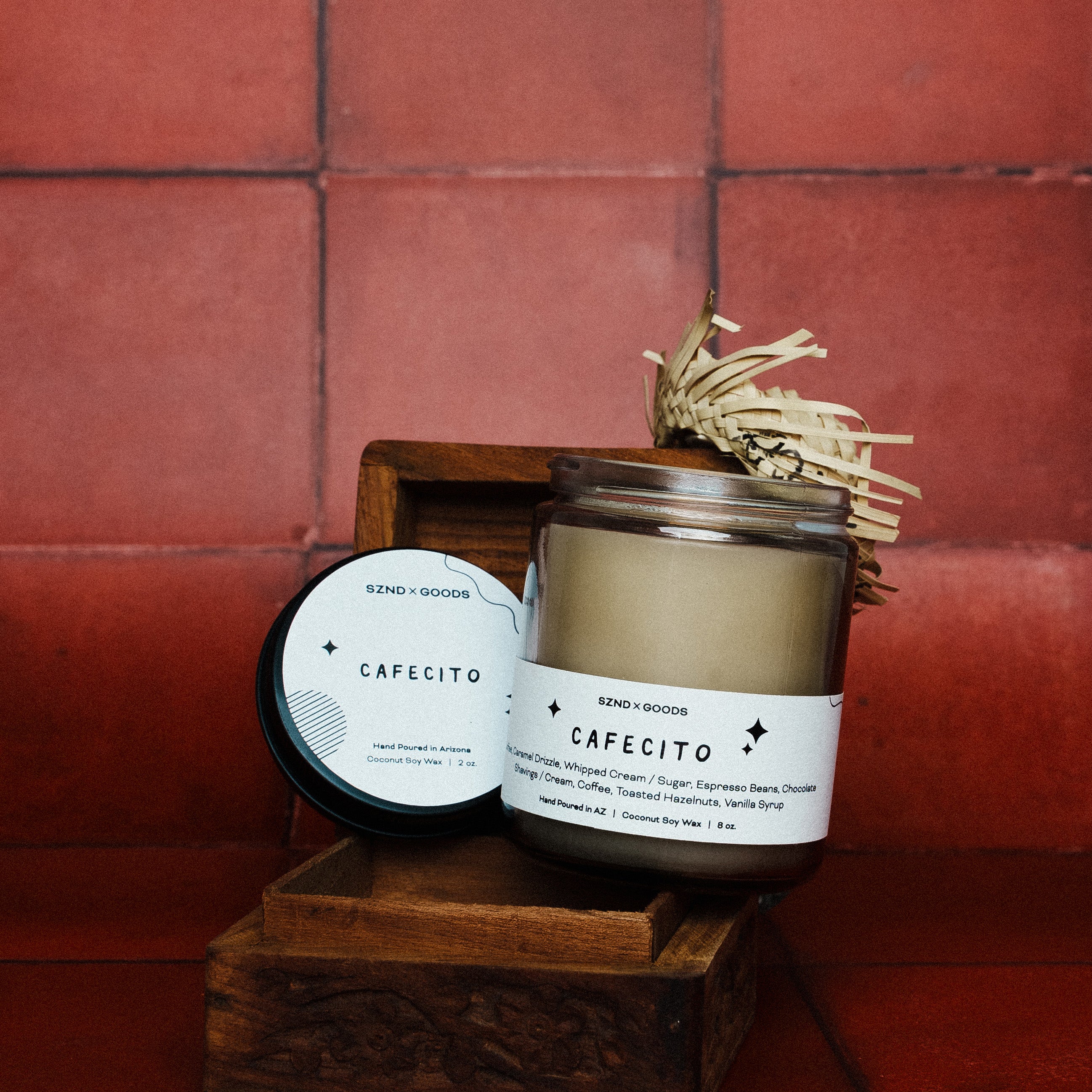 Cafécito candle with decorative straw on a wooden stand against a red tiled wall and a matching 2oz candle.