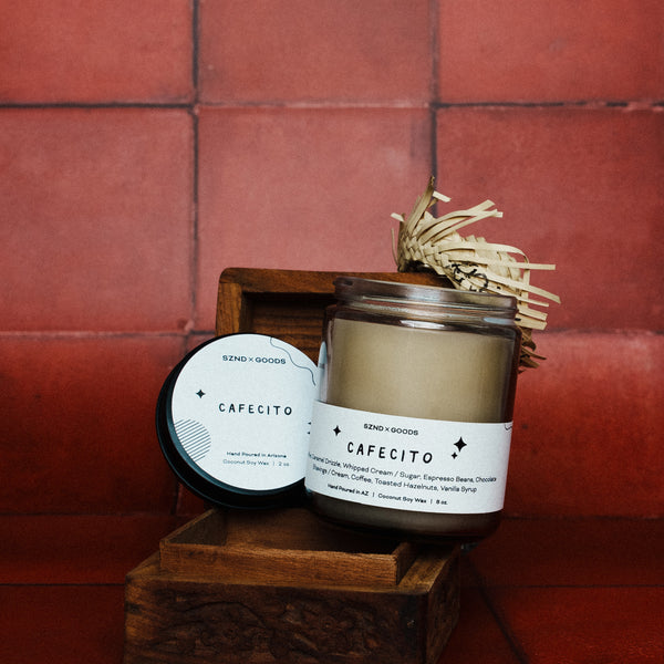 Cafécito candle with decorative straw on a wooden stand against a red tiled wall and a matching 2oz candle.