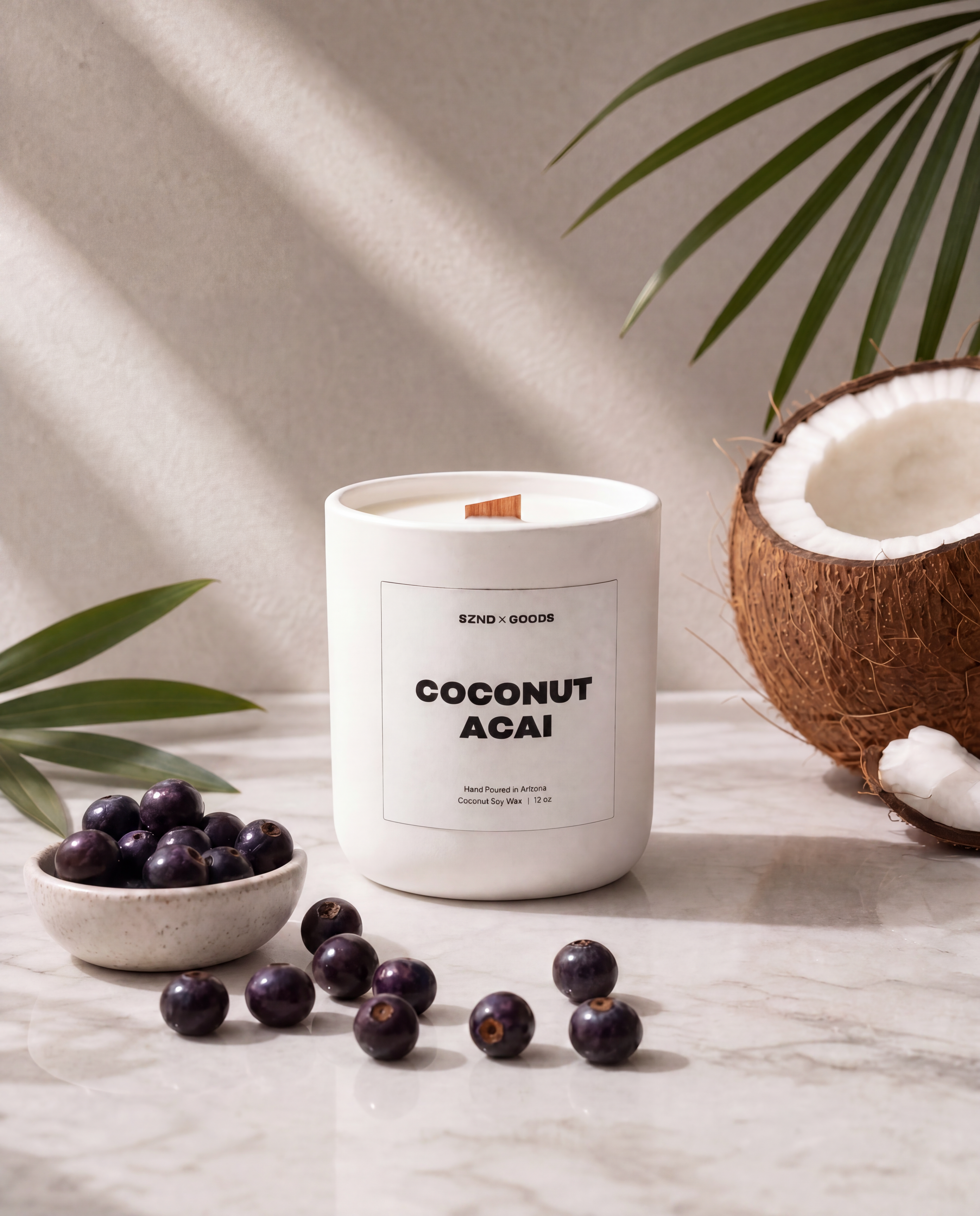 The Coconut Acai 12oz candle styled on a marble surface with acai berries, a halved coconut, and green palm leaves under soft diagonal shadows.