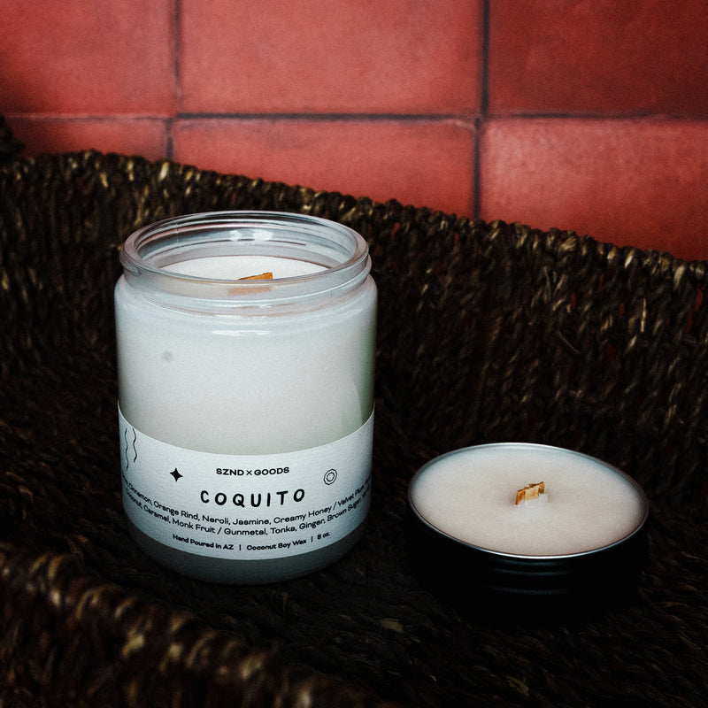 Candle labeled 'Coquito' on a woven surface with a red tiled wall background with it's matching mini 2oz candle.