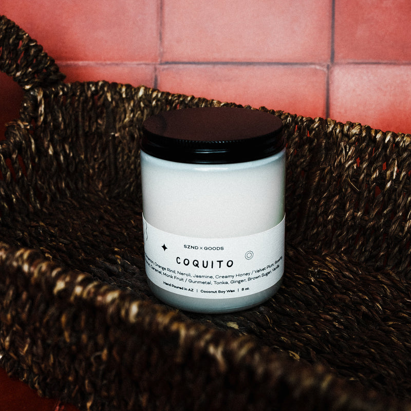 Candle labeled 'Coquito' in a woven basket with on a tiled wall background with candle lid.