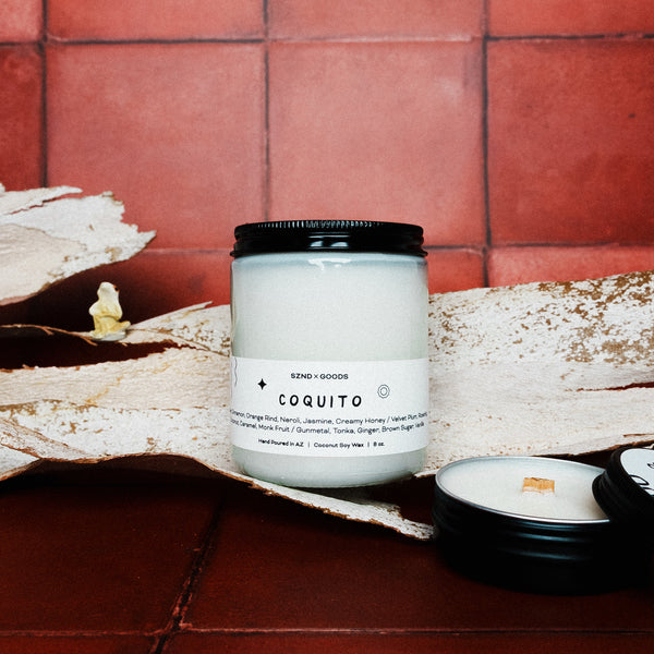 Candle labeled 'Coquito' on a rustic surface with a tiled wall background with 2oz candle and a coqui in the background.