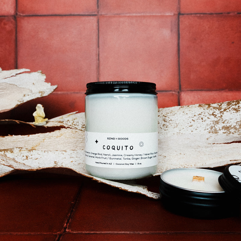 Candle labeled 'Coquito' on a rustic surface with a tiled wall background with 2oz candle and a coqui in the background.