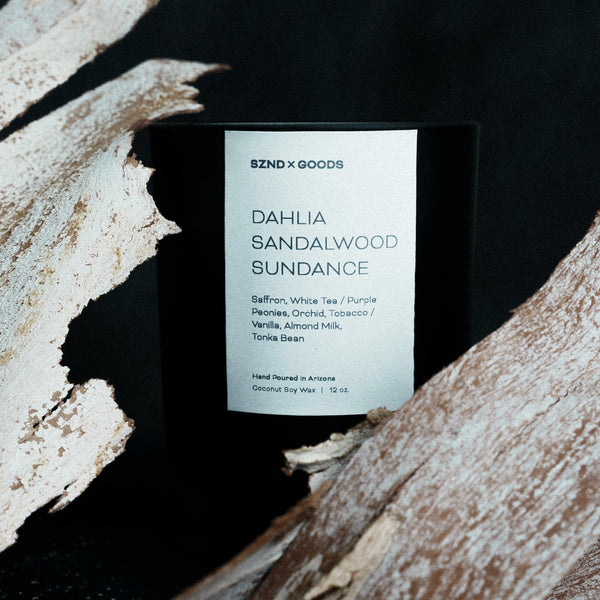 Candle labeled 'DAHLIA SANDALWOOD SUNDANCE' by SZND GOODS on a dark background with wooden elements.