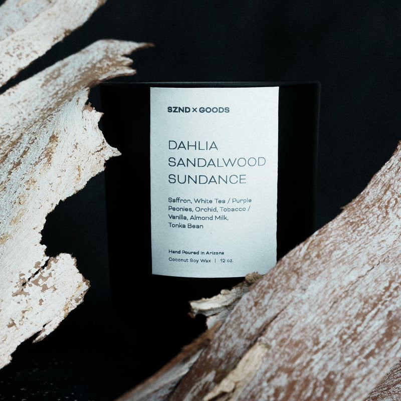 Candle labeled 'DAHLIA SANDALWOOD SUNDANCE' by SZND GOODS on a dark background with wooden elements.