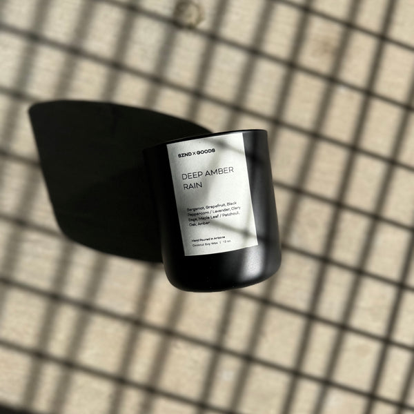 A black candle with a label that reads 'DEEP AMBER RAIN' is placed on a grid-patterned surface, casting a shadow over part of the grid.