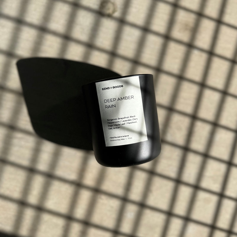A black candle with a label that reads 'DEEP AMBER RAIN' is placed on a grid-patterned surface, casting a shadow over part of the grid.
