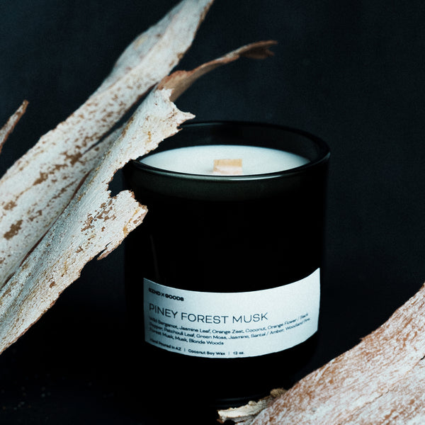 Wooden wick candle labeled 'Piney Forest Musk' in a black container on a dark background with natural elements. 