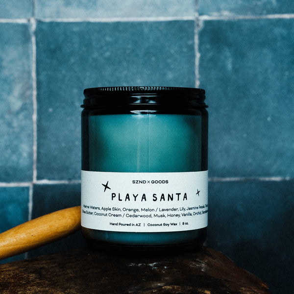 Candle jar labeled 'Playa Santa' with a wooden stick on a blue tiled background