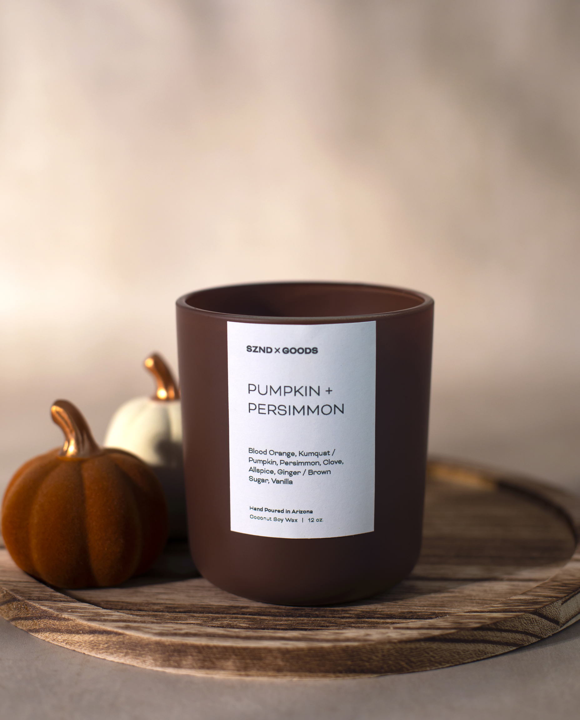 A brown candle jar with a white label reading Pumpkin + Persimmon styled on a wooden tray with small pumpkins against a soft beige background.