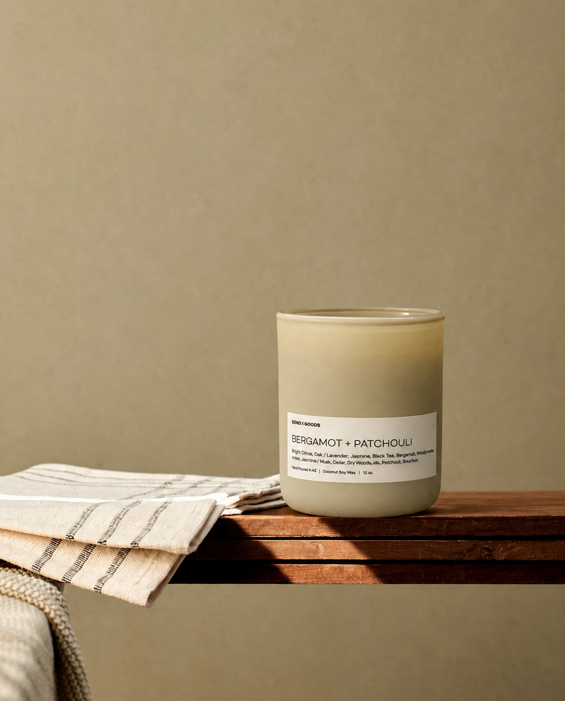 Our Bergamot + Patchouli candle in a matte cream-colored glass jar, styled on a wooden ledge with a folded striped cloth against a warm beige background.