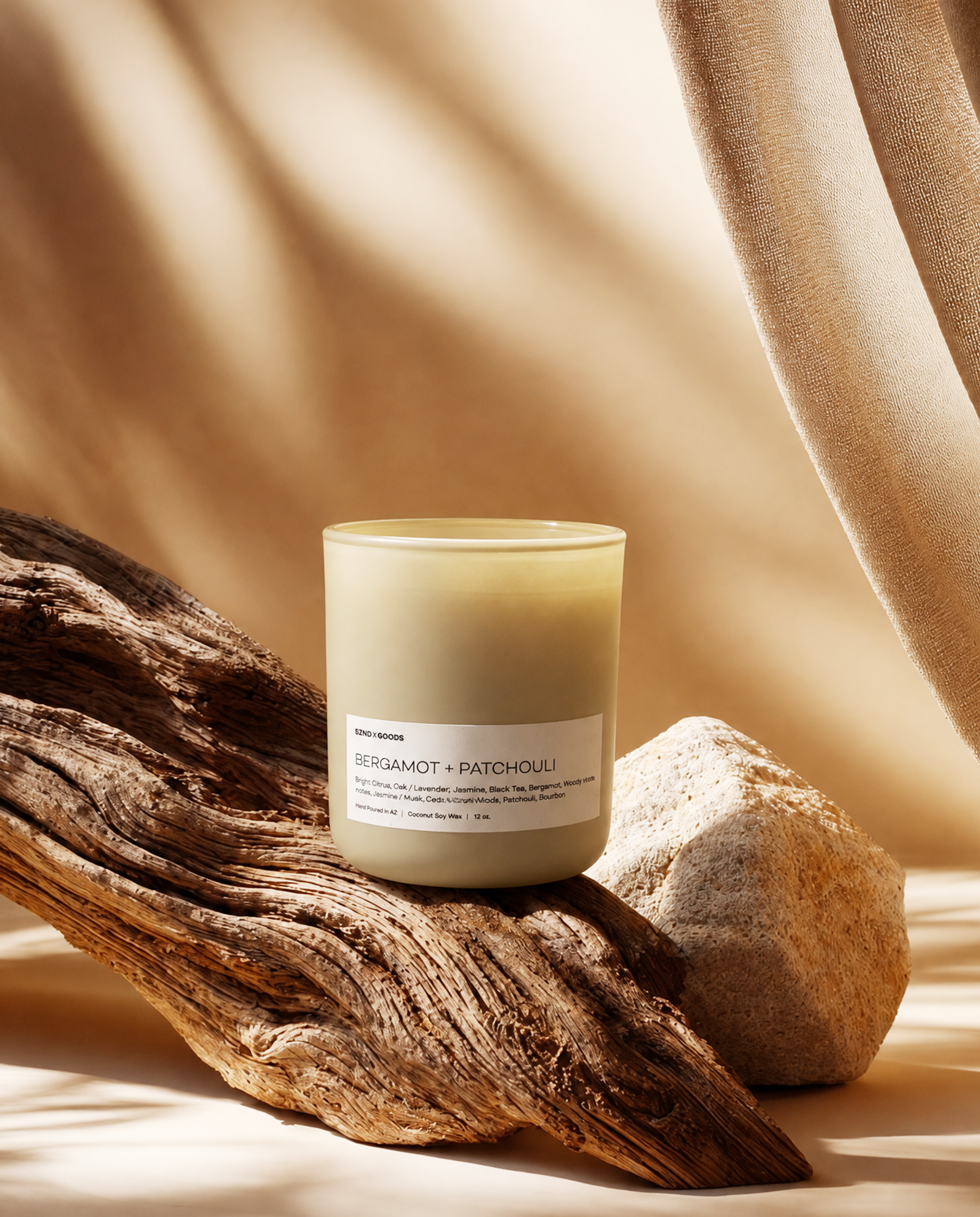 A Bergamot + Patchouli glass jar candle with creamy off-white wax sits on a piece of weathered driftwood next to a stone, styled with draped beige fabric in warm, sunlit shadows. 
