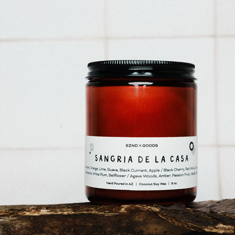 Red candle jar labeled 'Sangria de la Casa' on a wooden surface with a white tiled background