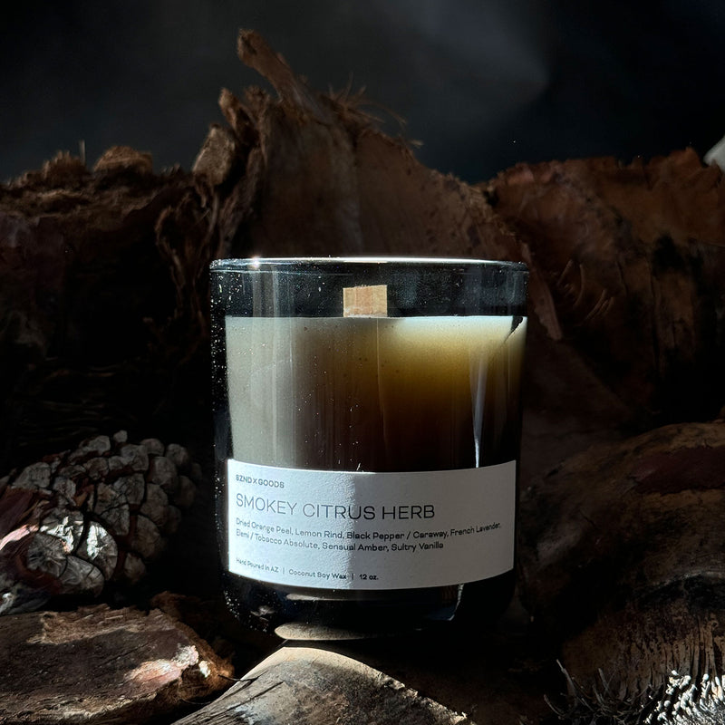 A brown glass candle with a wooden wick, labeled 'Smokey Citrus Herb', placed on a rustic wooden surface with pine cones and bark in the background.