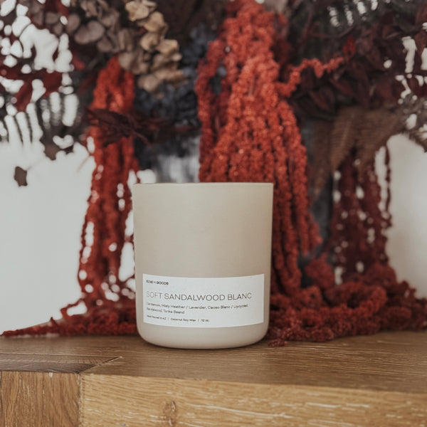 A beige 12oz candle with a wooden wick, labeled 'Soft Sandalwood Blanc', placed on a wooden surface with a red textured object in the background.