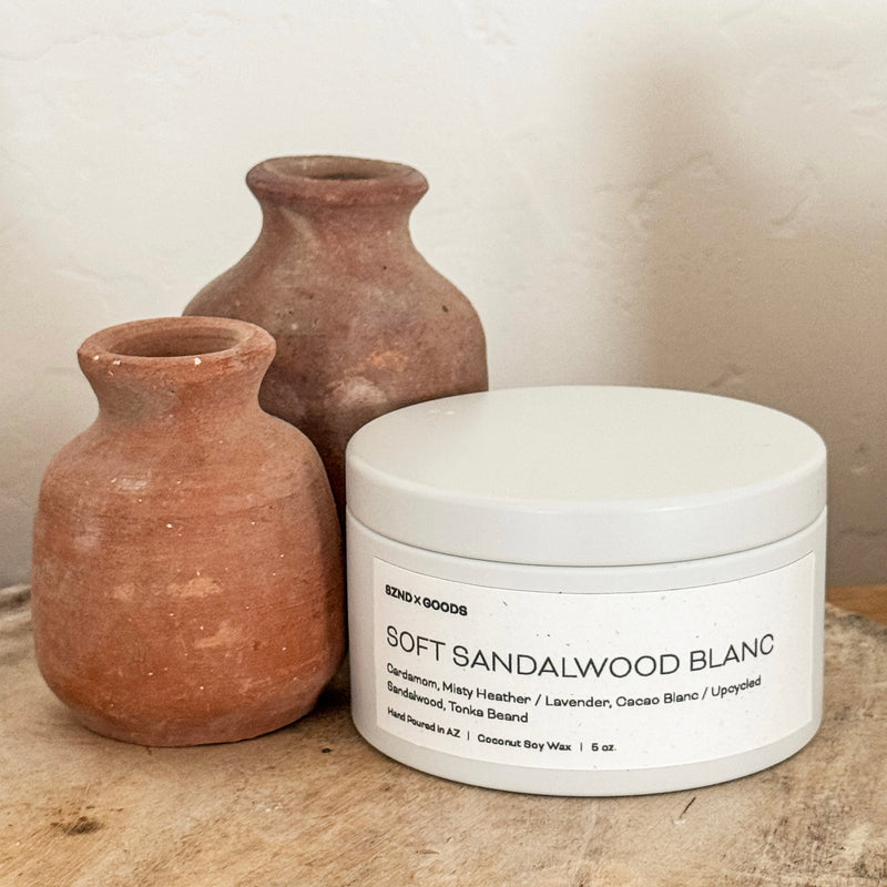 White candle container labeled 'Soft Sandalwood Blanc' with two terracotta vases on a wooden surface.