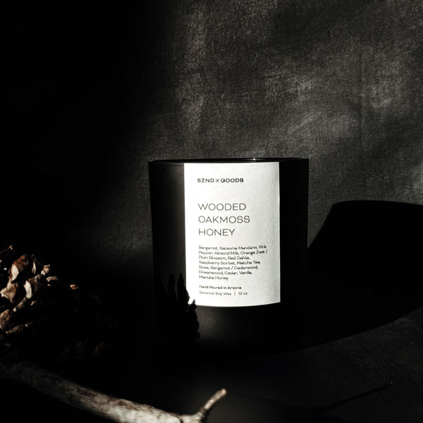A black candle with 'Wooded Oakmoss Honey' text on its label, placed on a dark surface with a pine branch in the foreground.