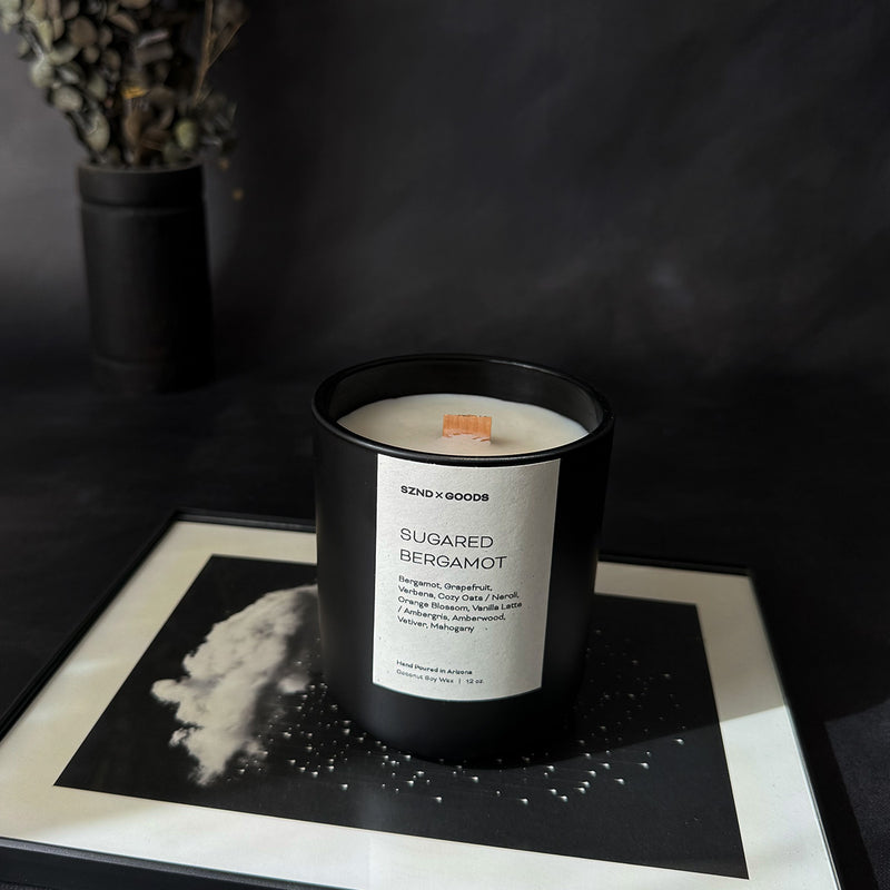 A black container candle with a wooden wick, labeled 'SUGARED BERGAMOT', with a white wax melt beside it, on a dark background.