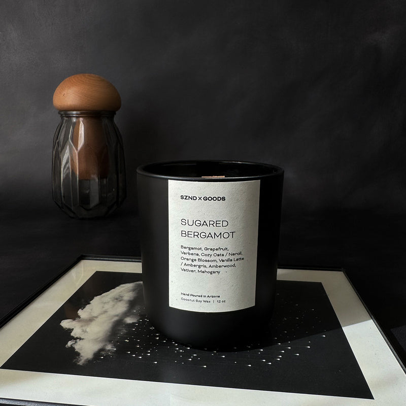 A black container candle with a wooden wick, labeled 'SUGARED BERGAMOT', with a white wax melt beside it, on a dark background.