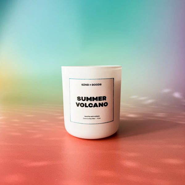 A white candle with a wooden lid labeled 'SUMMER VOLCANO' on a dual-tone background with blue at the top and pink at the bottom.