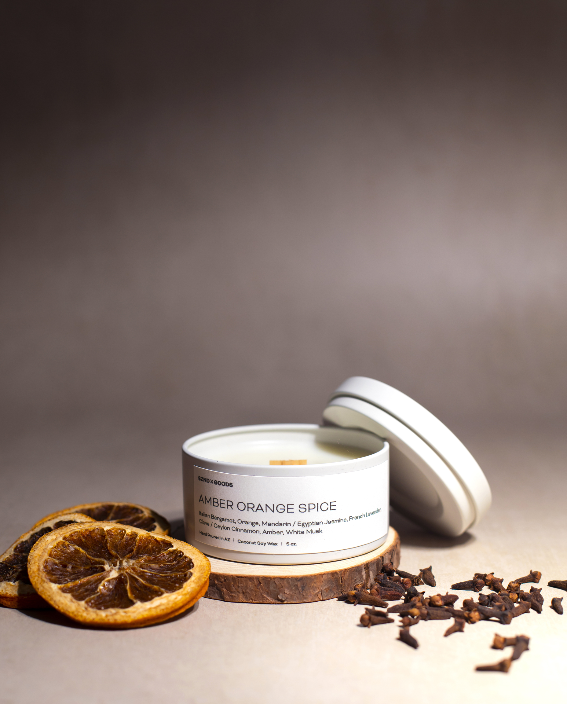 A white metal tin candle labeled 'AMBER ORANGE SPICE' sits on a wooden coaster with dried orange slices and cloves around it on a warm taupe surface against a dark brown gradient background.