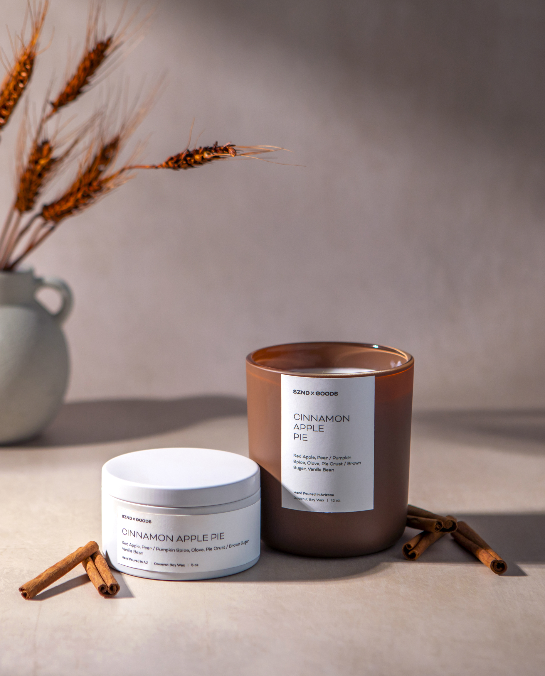 Our Cinnamon Apple Pie candle styled on a beige surface with scattered cinnamon sticks and dried wheat in a vase against a neutral background.