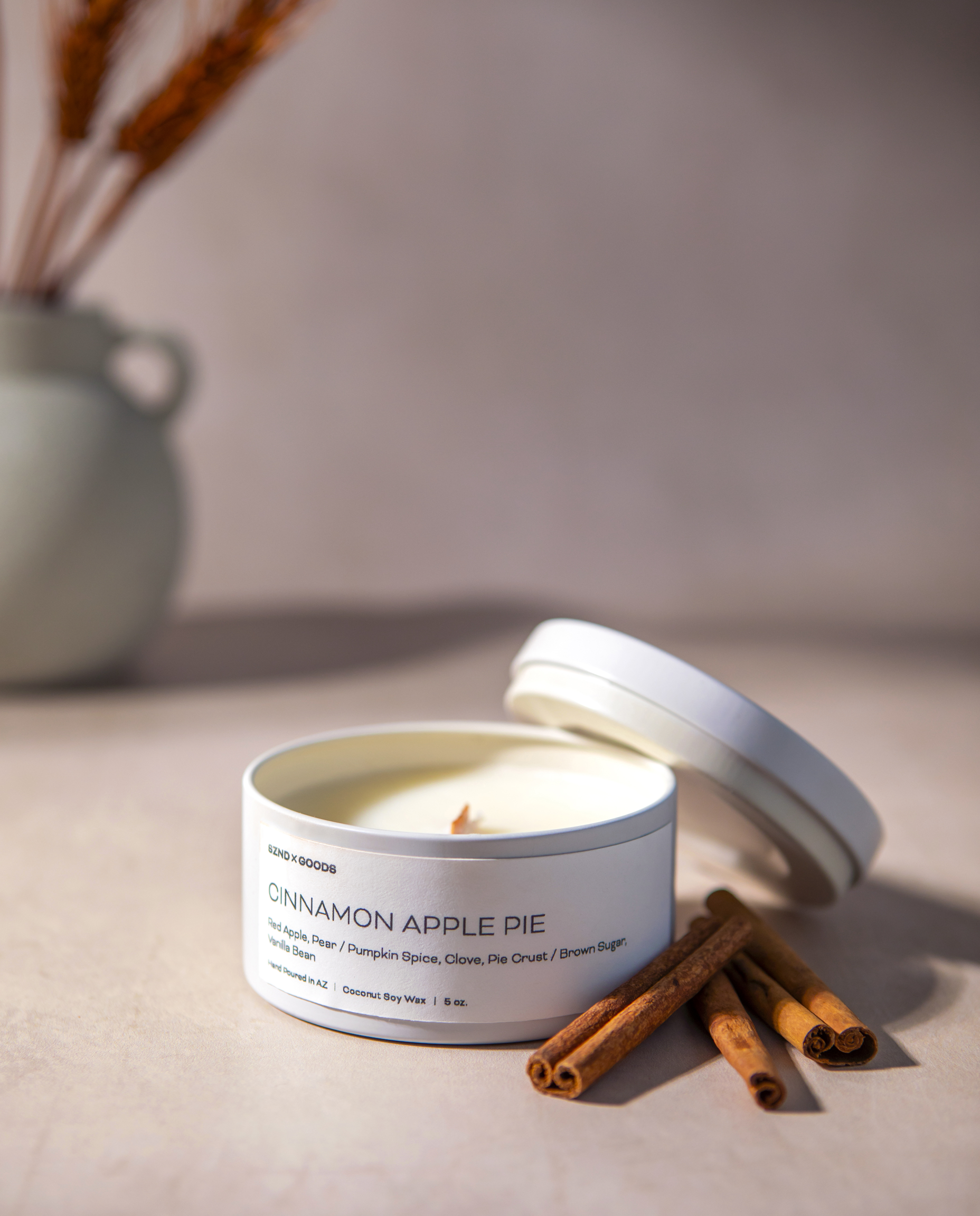 The Cinnamon Apple Pie 5oz shown open with its lid leaning against it on a beige surface. Several cinnamon sticks sit beside the tin, and a softly blurred ceramic vase with dried stems appears in the background.