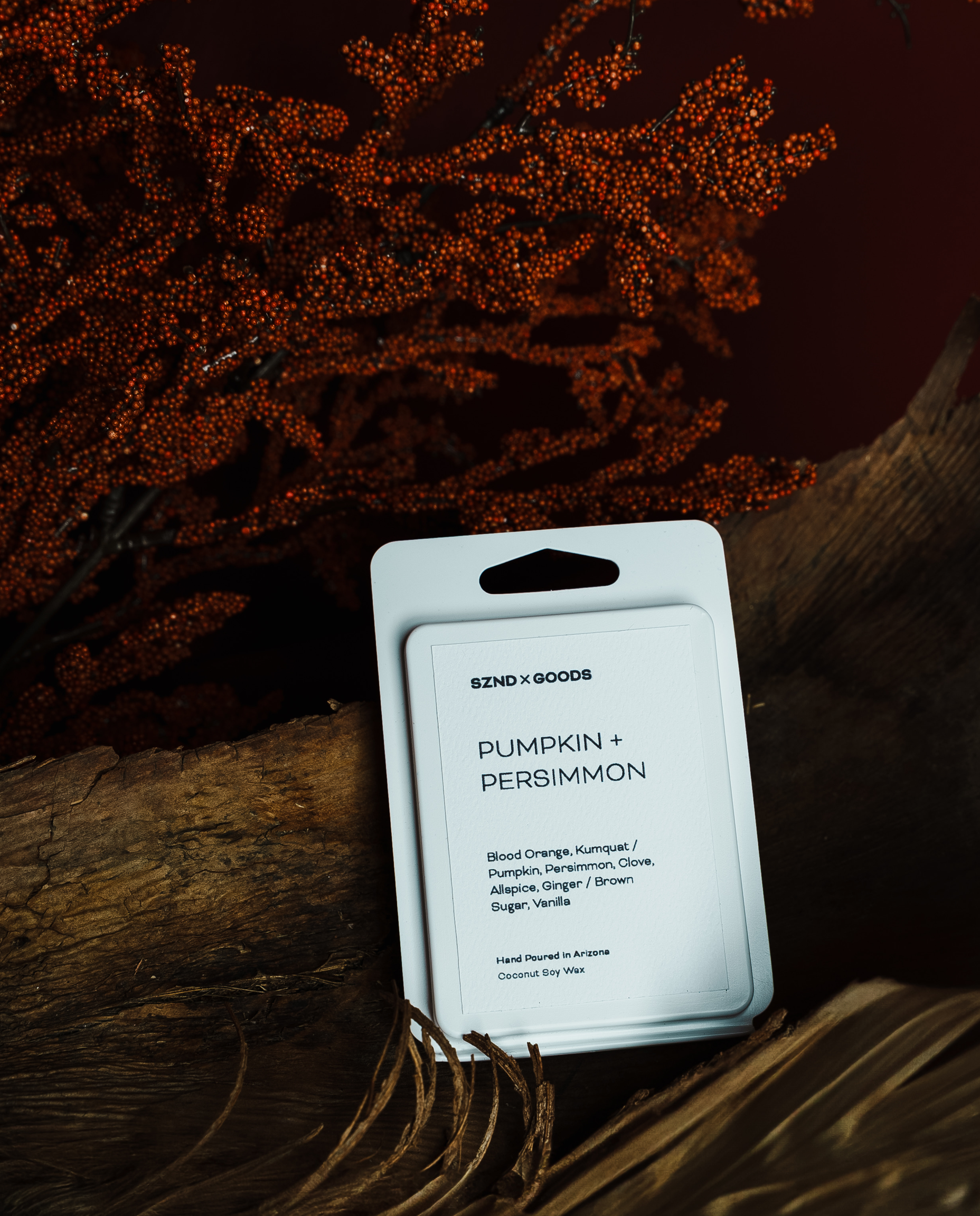 A packaged fragrance wax melt card labeled 'PUMPKIN + PERSIMMON', placed on a rustic wooden surface with dried orange-brown botanicals in the background.