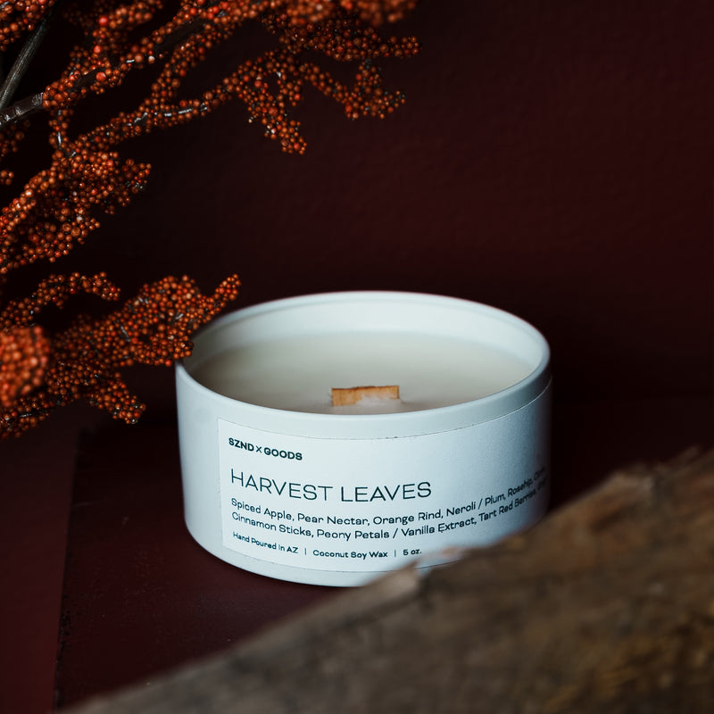 Wooden wick candle in a white container labeled 'Harvest Leaves' with decorative elements in the background.