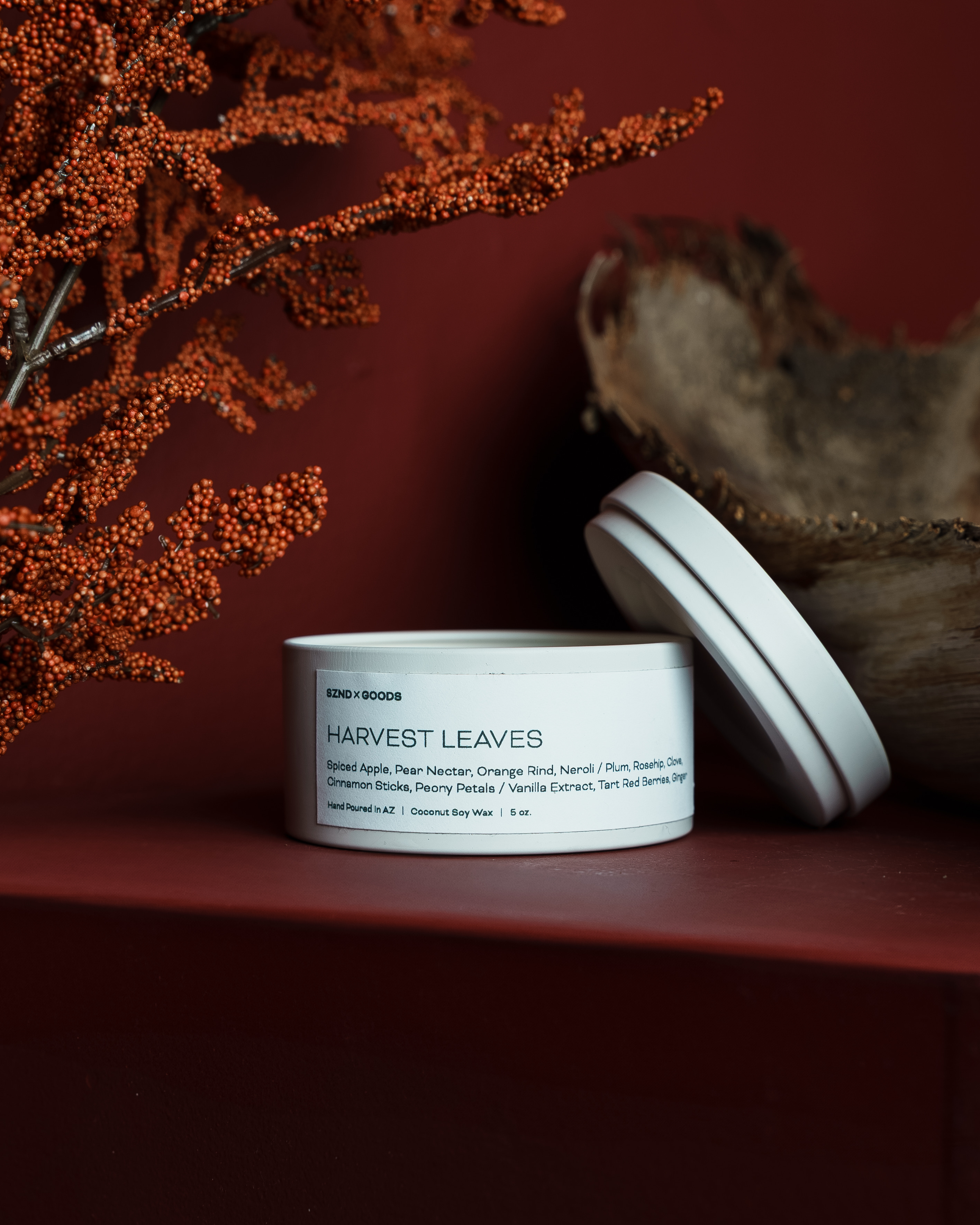A white metal tin of 'Harvest Leaves' scented candle, shown with its lid leaning against it on a dark red surface. The background features a dark red wall, dried orange berries, and a piece of driftwood.