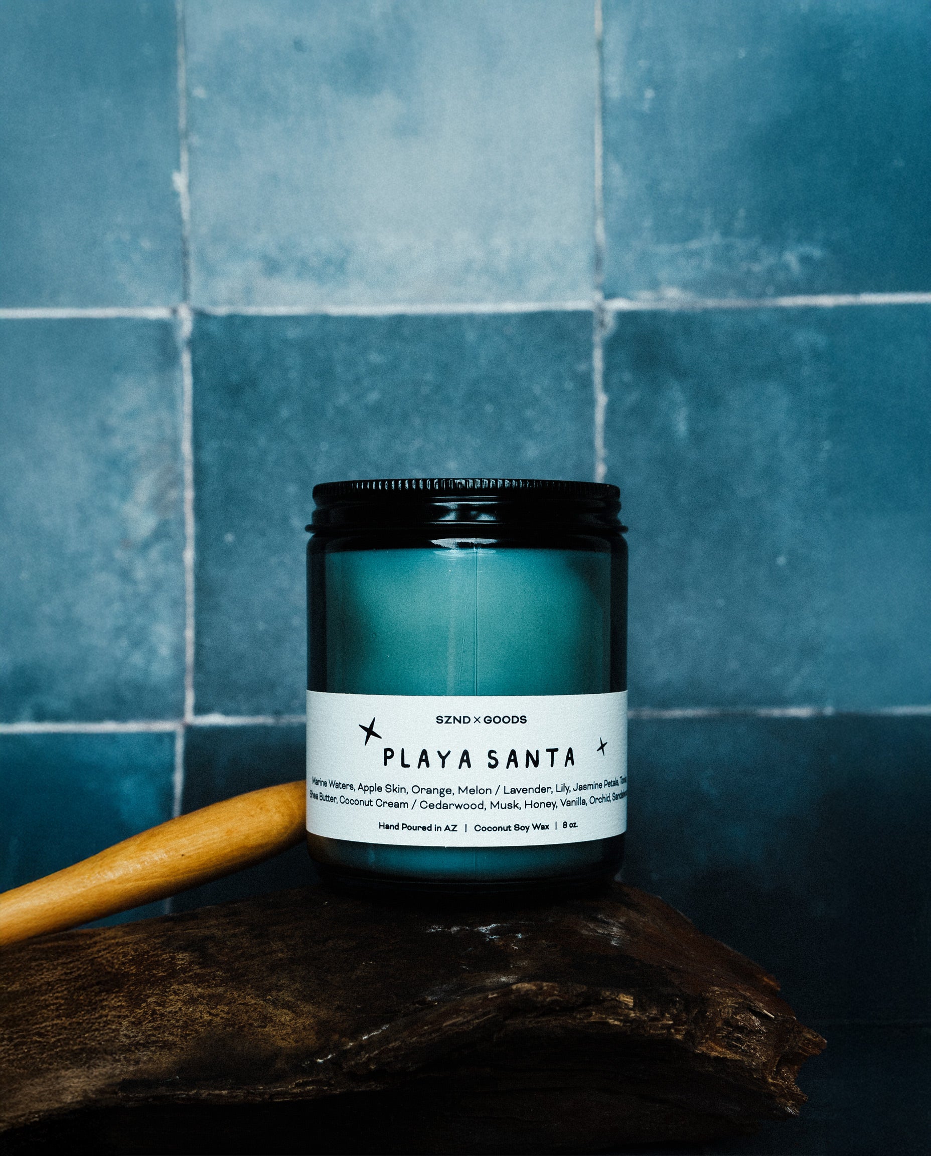 A Playa Santa wodoen wick candle sits on a piece of dark wood against a blue tiled background. A wooden spoon rests behind the candle.