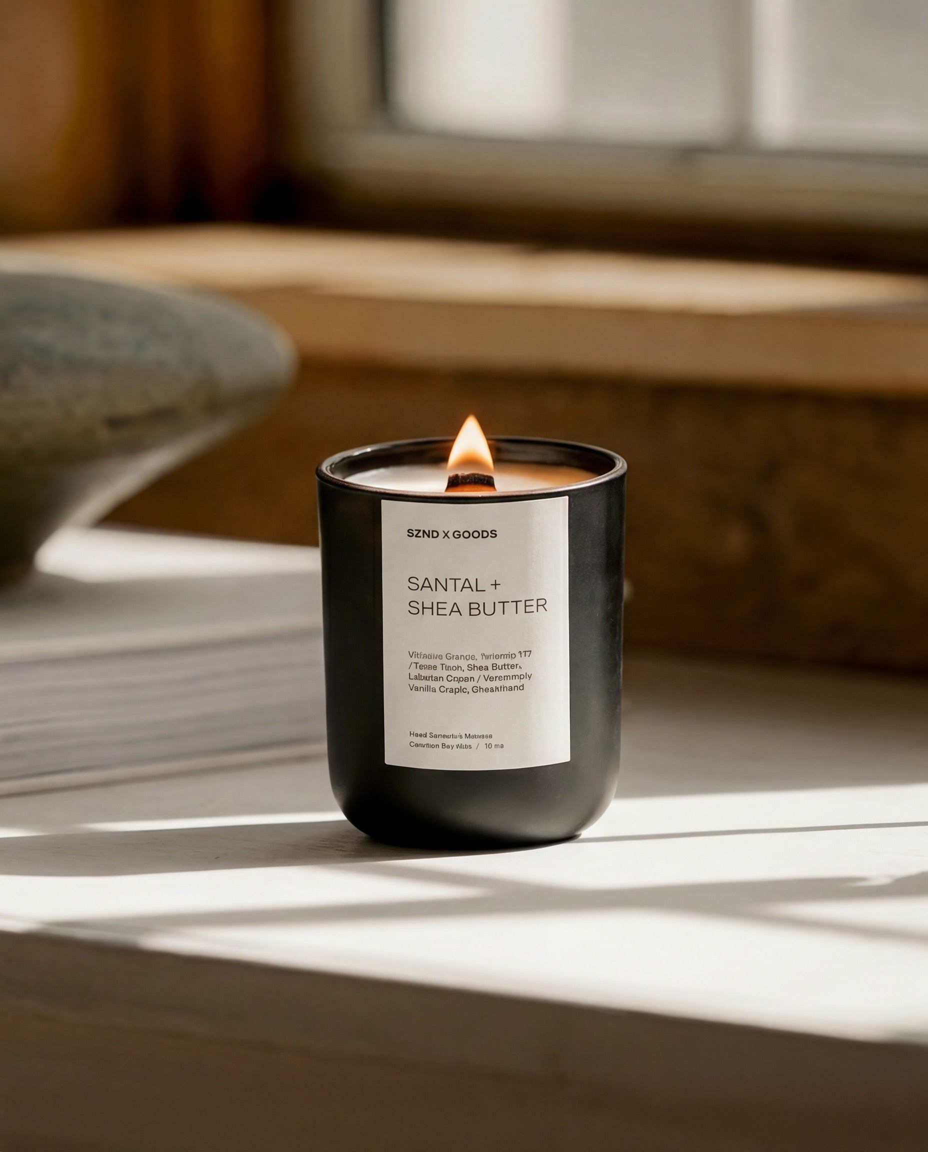 A black glass jar candle with a white label reading 'SANTAL + SHEA BUTTER' is lit on a windowsill with warm sunlight and soft shadows.