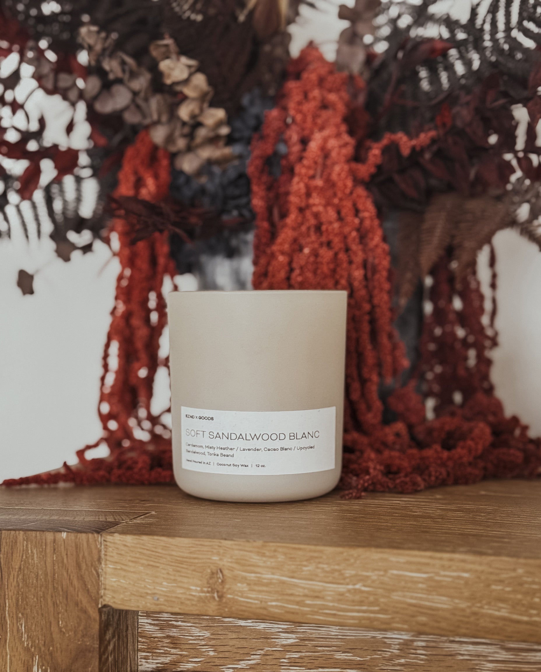 A coconut soy candle in a light cream jar sits on a rustic wooden table, styled with dried ferns and rust-red botanicals in the background.