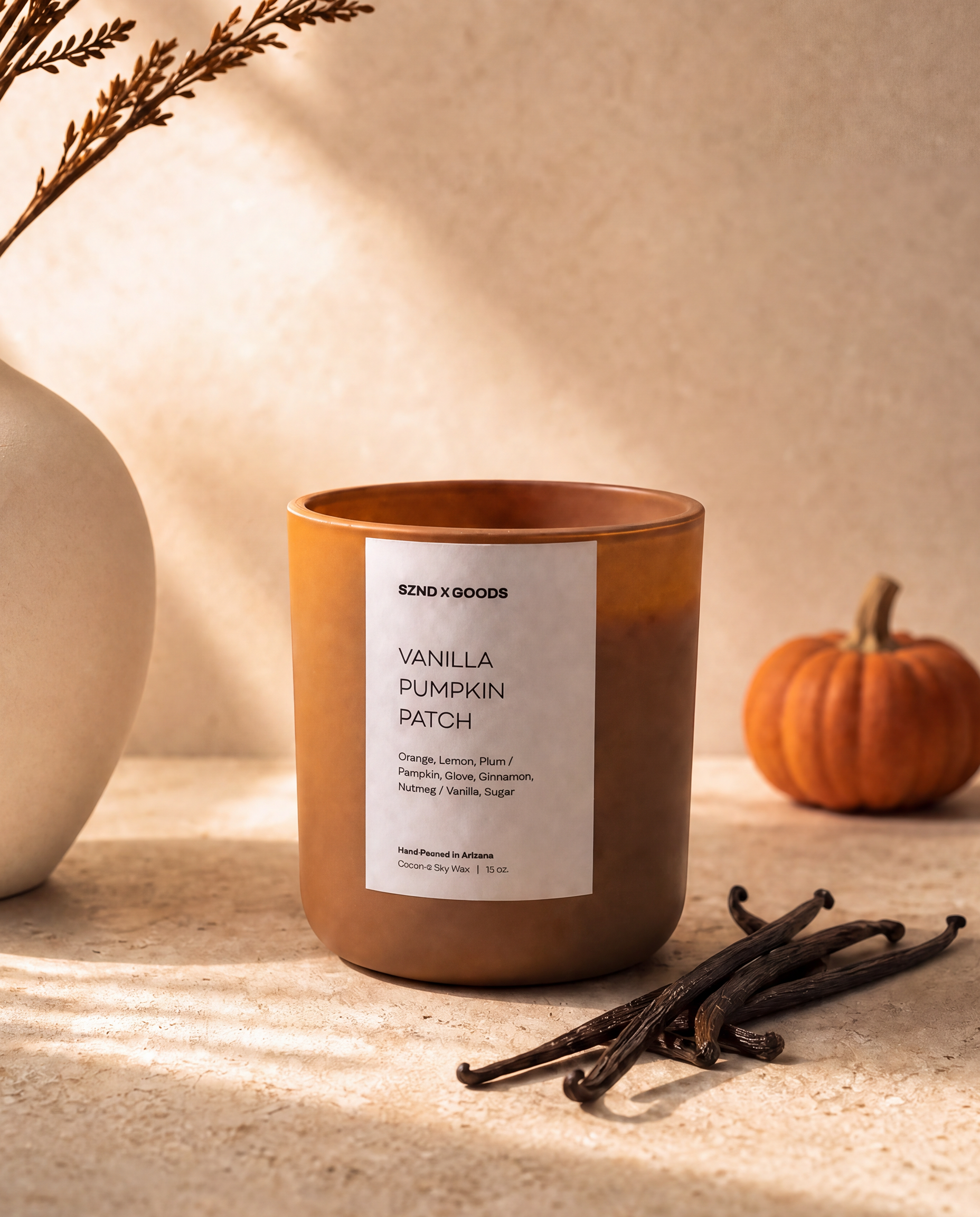 A candle in a brown glass jar with a white label reading Vanilla Pumpkin Patch, styled on a beige surface with vanilla beans, a small pumpkin, and a ceramic vase in warm sunlight.