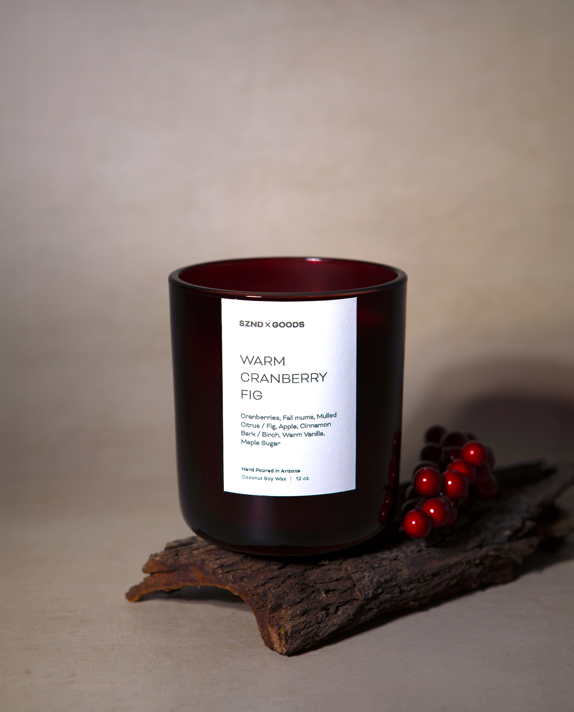 A dark amber glass candle jar with red cranberry berries on a rustic wood prop against a neutral beige background.
