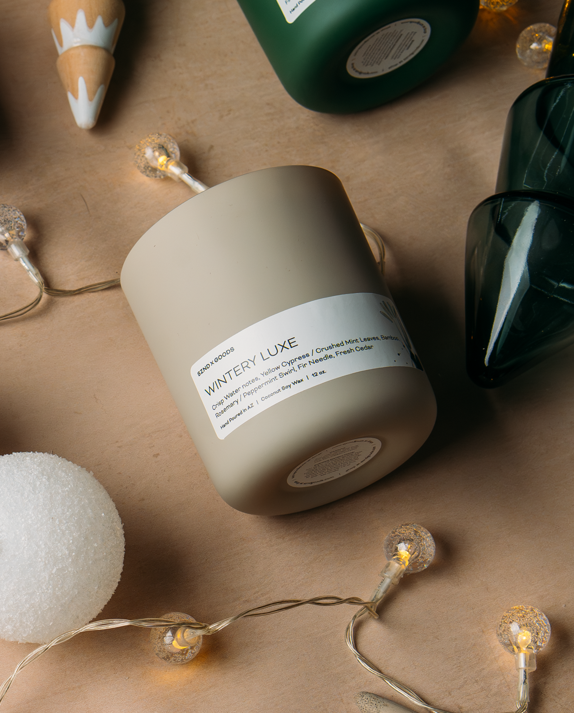 A matte gray candle jar labeled 'WINTERY LUXE' by SZND GOODS, styled on a brown kraft paper background with warm fairy lights, a white pom-pom ball, and dark green glass decor.