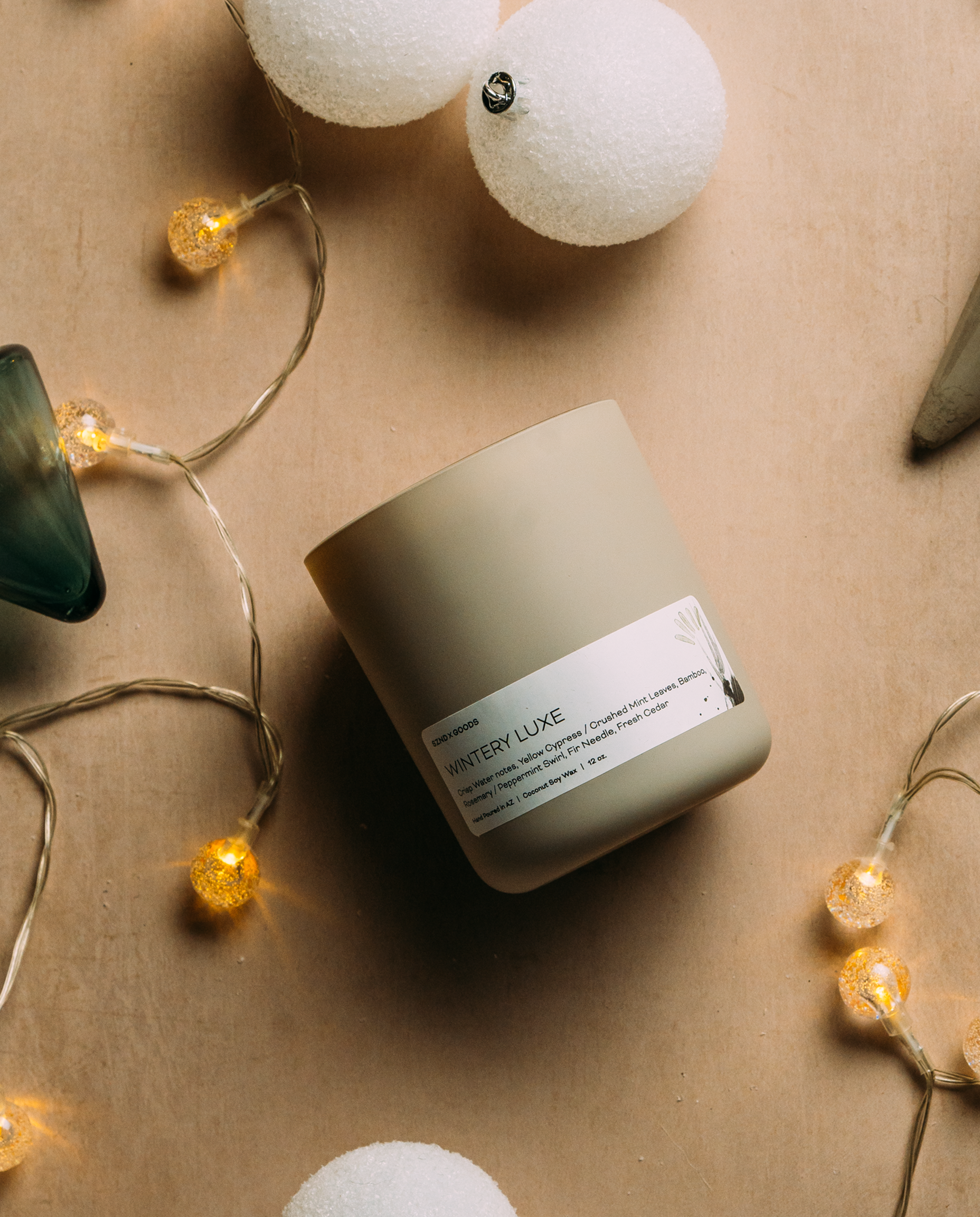 A matte gray candle jar with a white label reading 'Wintery Luxe' by SZND GOODS, styled on a beige background with warm fairy lights, white pom-pom ornaments, and a dark green glass object.